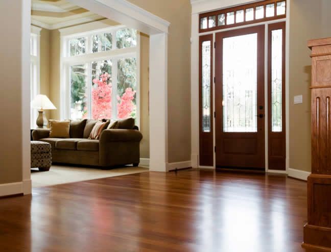 Bright empty living room with hardwood floors, large front windows, and a glass-paneled wooden door