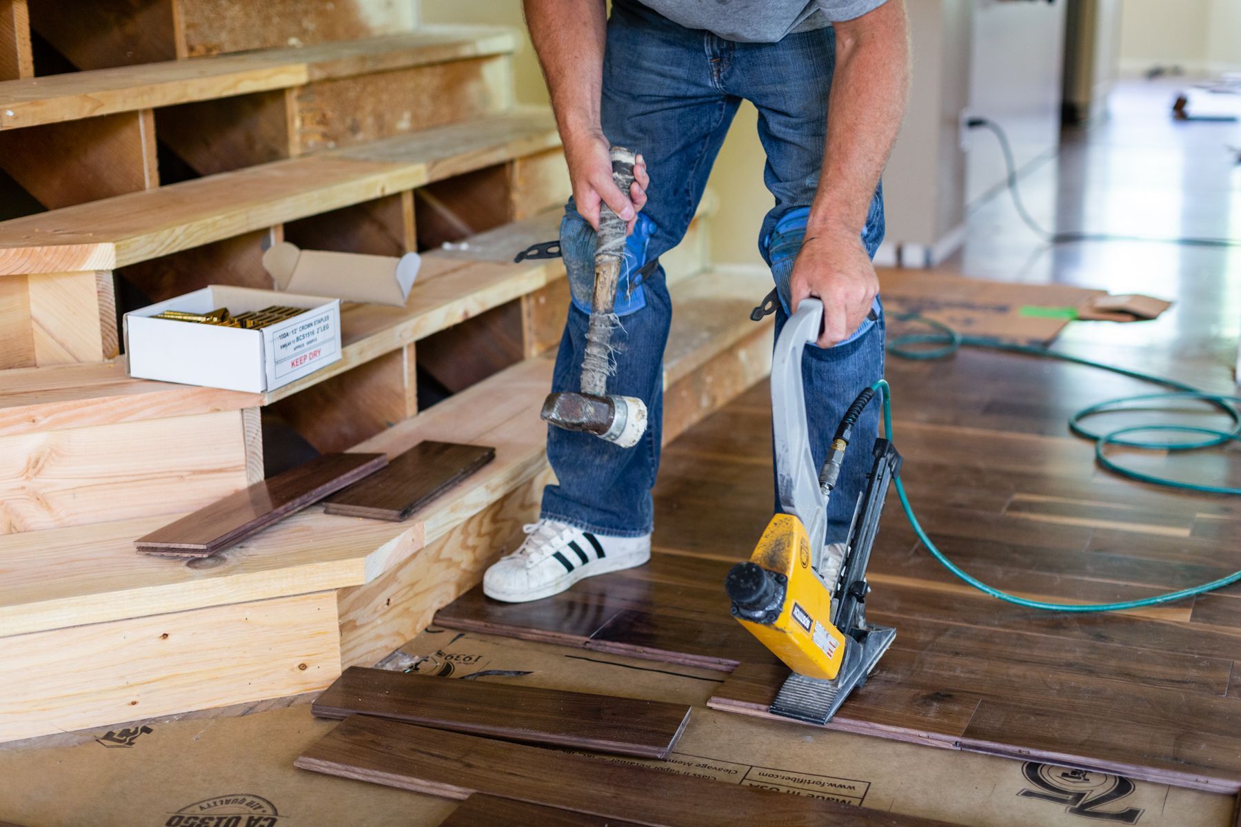 Worker kneeling on plywood subfloor, using a nail gun to install wooden steps at a construction site