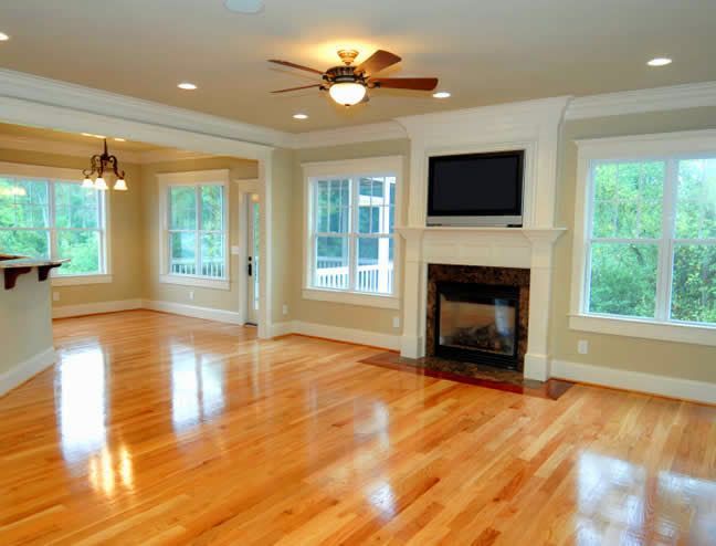 Bright empty living room with hardwood floors, large windows, ceiling fan, and fireplace with TV above it