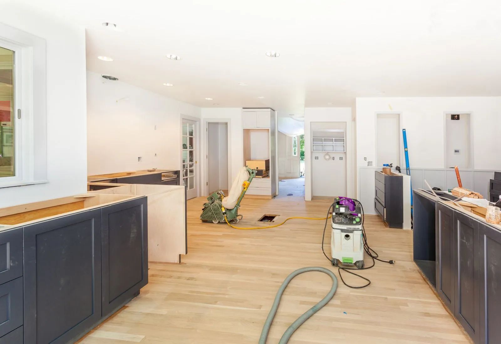 Bright kitchen under renovation with dark cabinets, wood floors, tools, and a vacuum on the floor