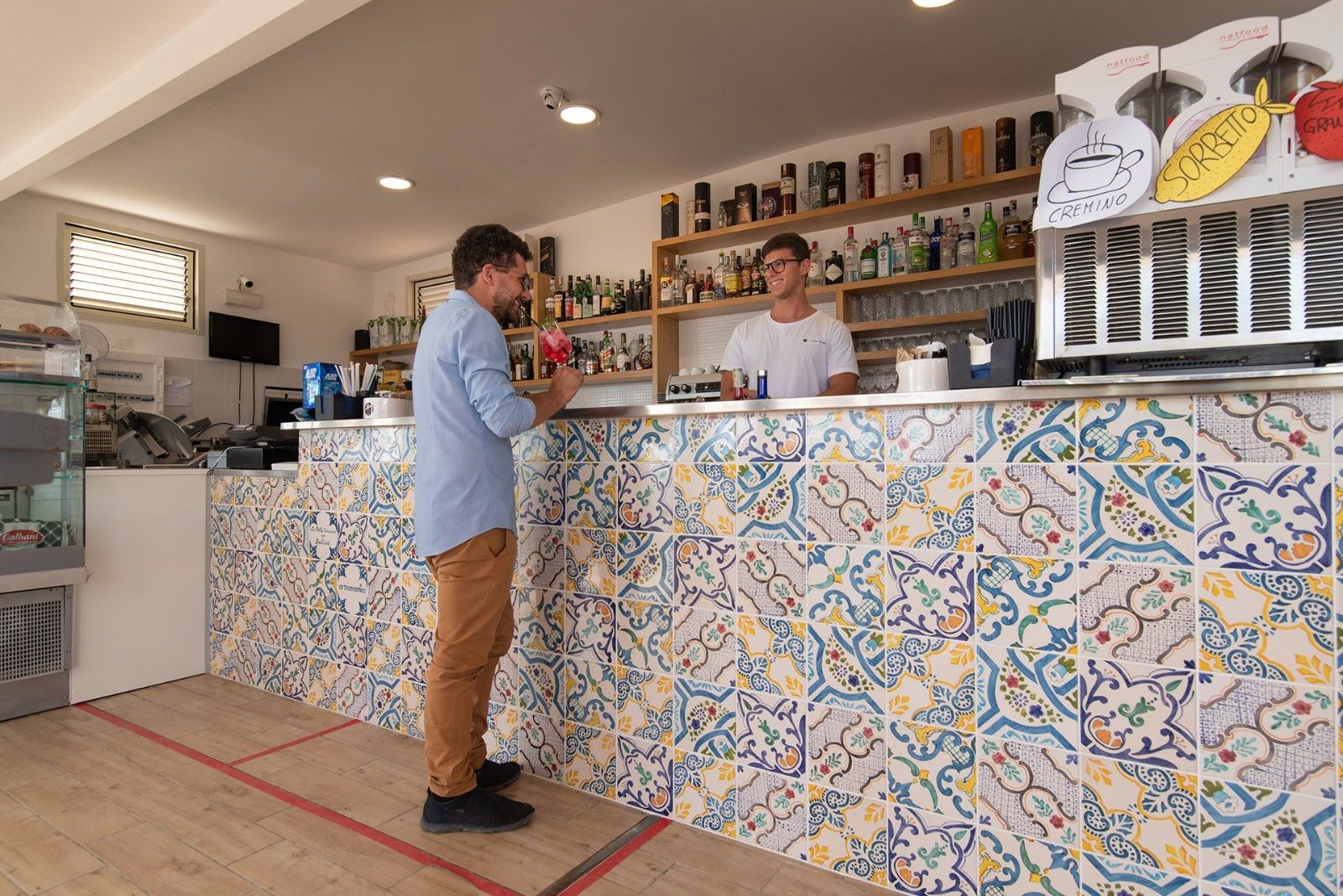 Due persone che chiacchierano al bancone di un caffè con piastrelle colorate, dietro al quale si trovano bottiglie e menù.