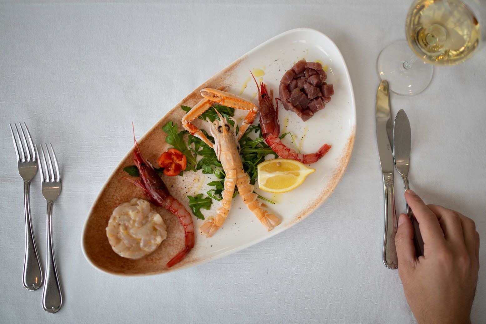 Piatto di frutti di mare, limone e un bicchiere di vino bianco accanto a una mano che regge coltello e forchetta.