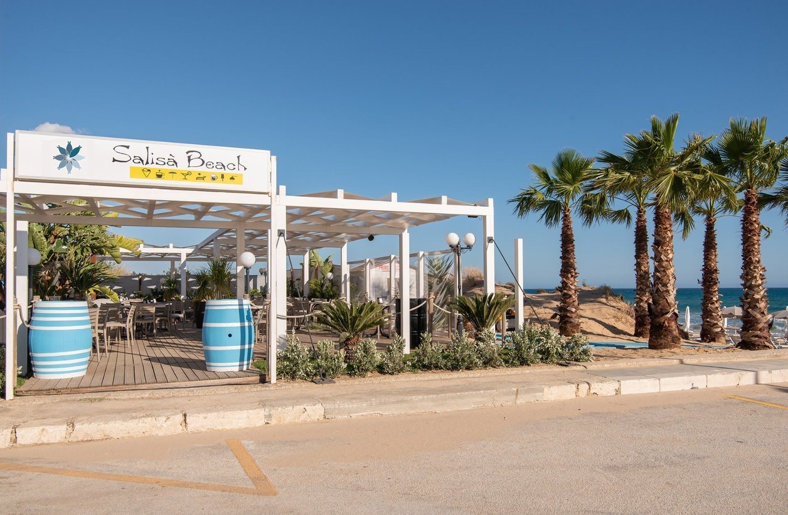 Beachfront café with white pergola, blue barrels, and palm trees by the road