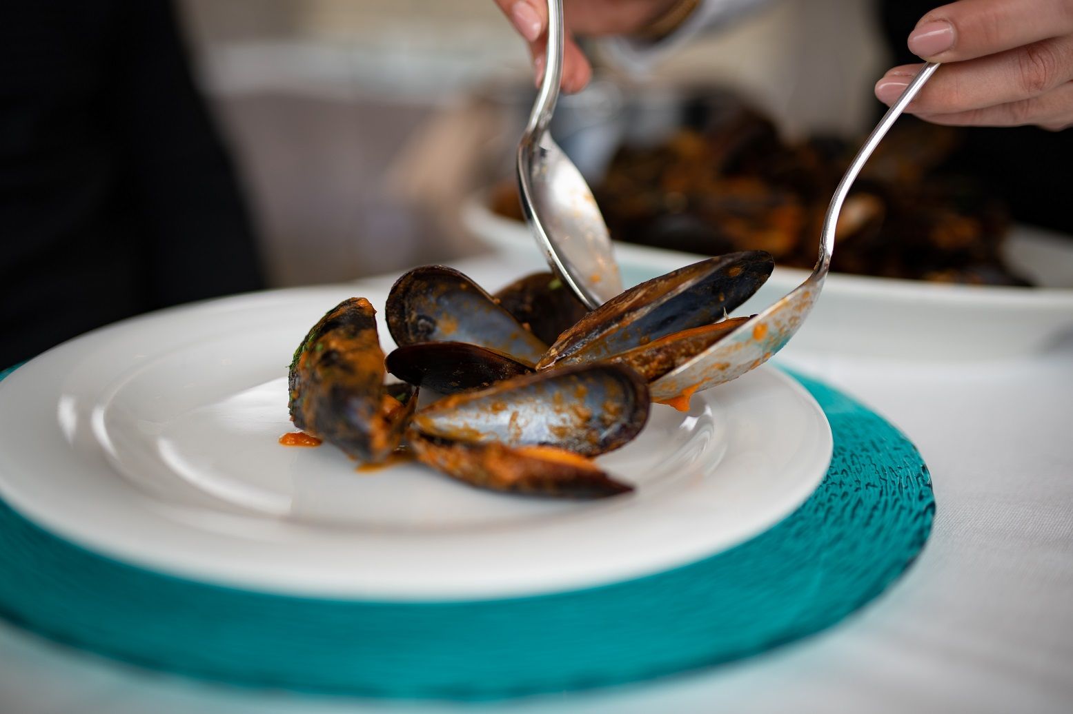 Cozze su un piatto bianco con una tovaglietta color verde acqua, servite con pinze.