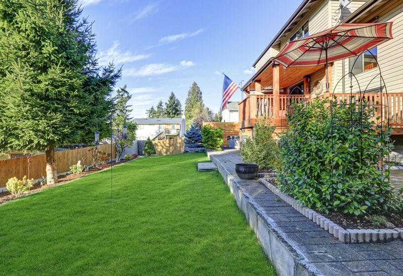 Backyard with green lawn, wooden deck, flag, and trees under a sunny blue sky.