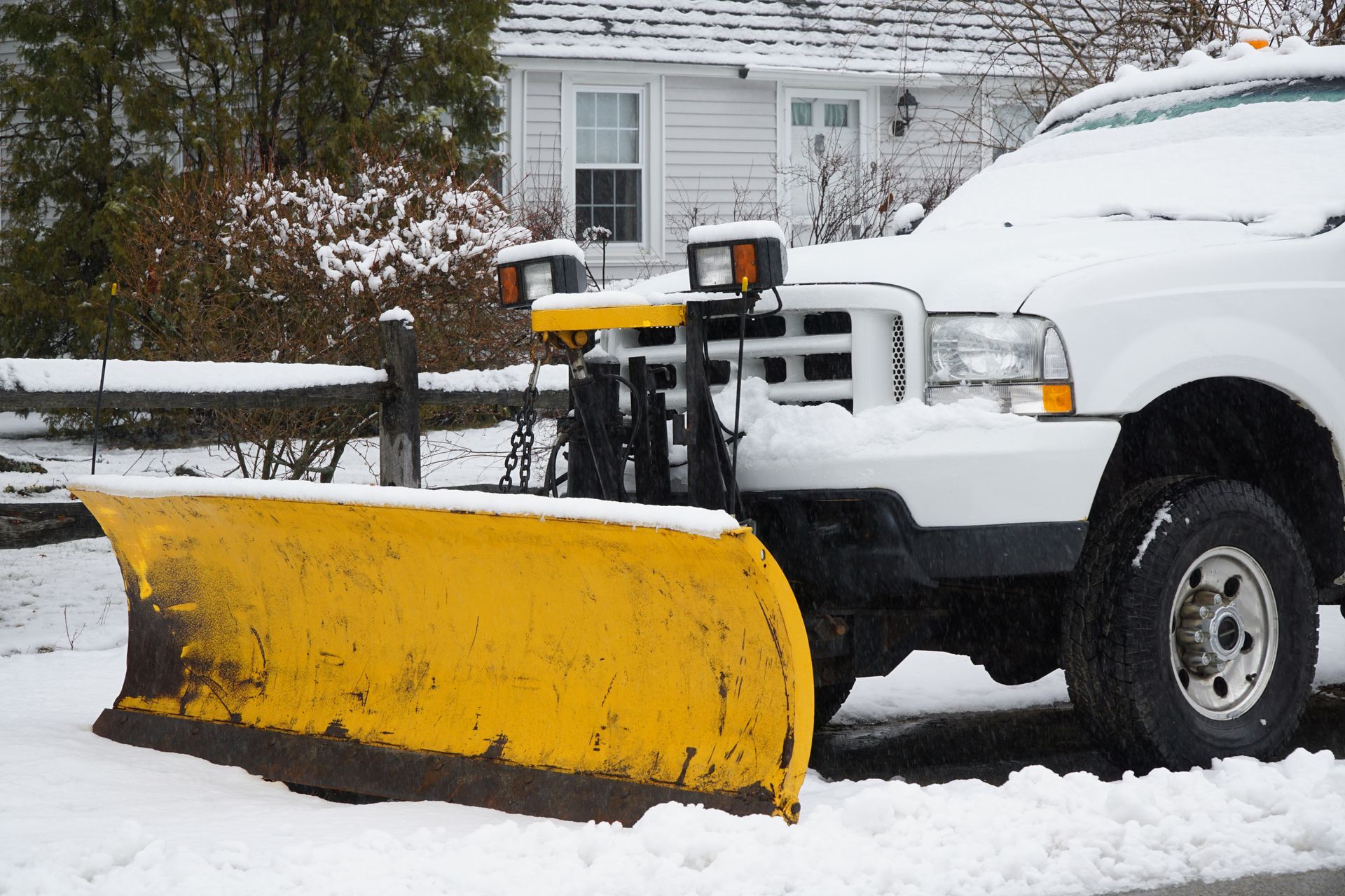 Yellow snowplow on a white truck clearing snow in front of a house.