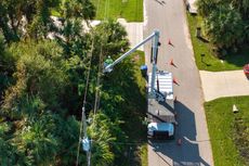 Aerial view of a utility truck with bucket arm on a tree-lined street, with orange cones nearby.