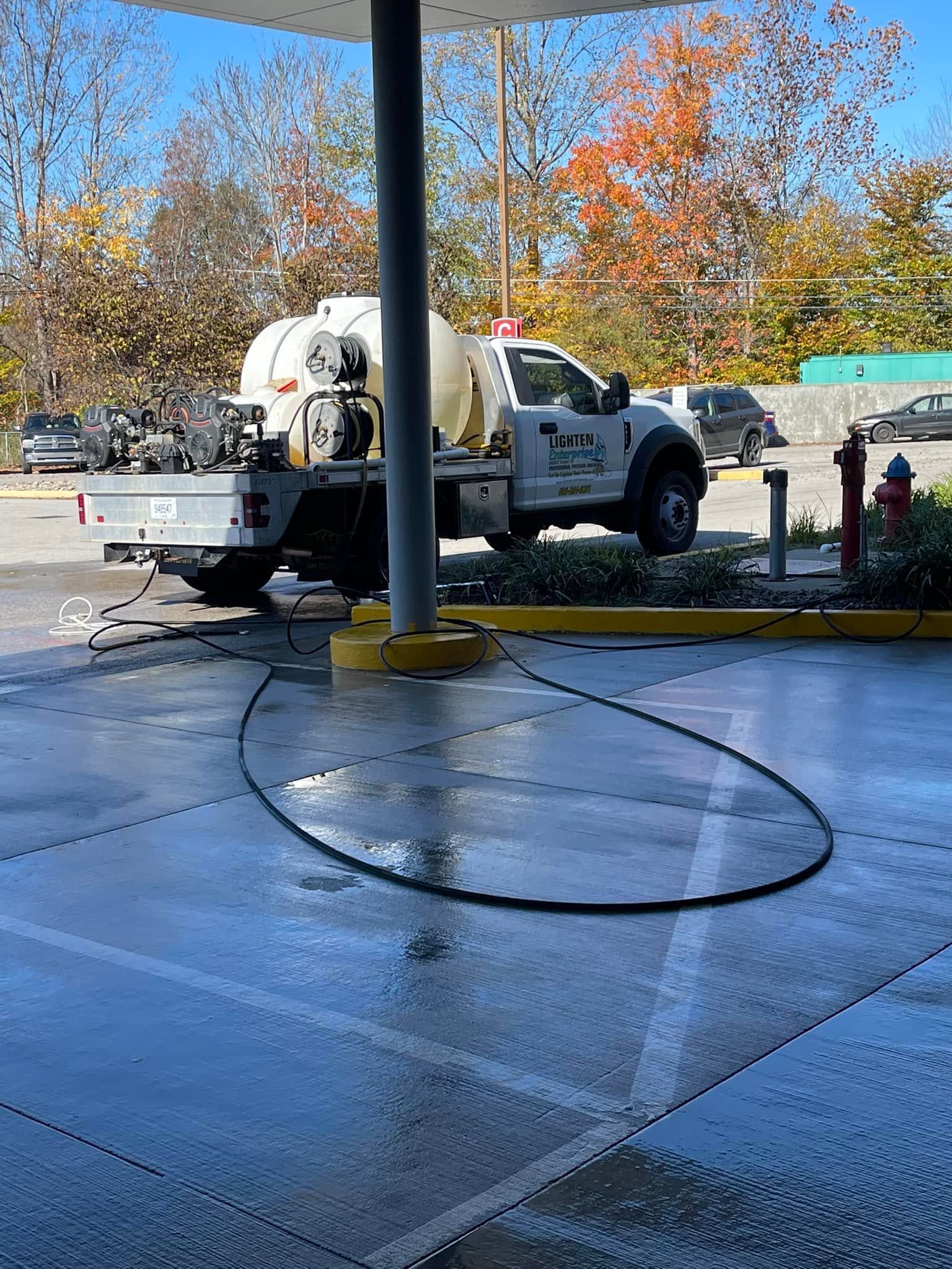 White utility truck parked by a gas station pump, with a hose looped across wet pavement under a blue sky.