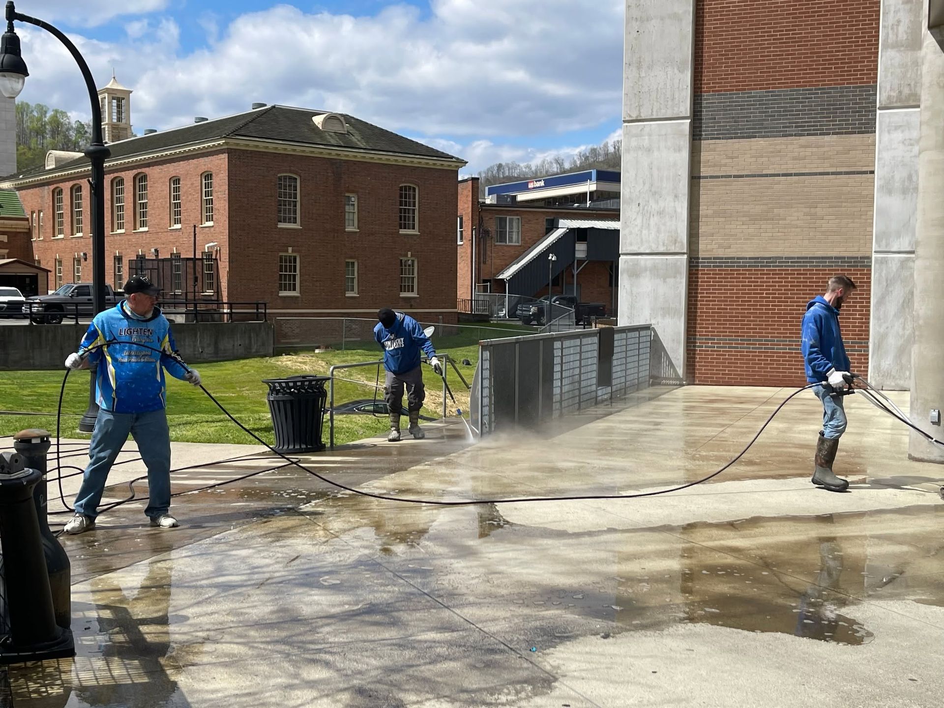 Workers washing pavement outdoors beside a brick building and puddles, with hoses spraying water.
