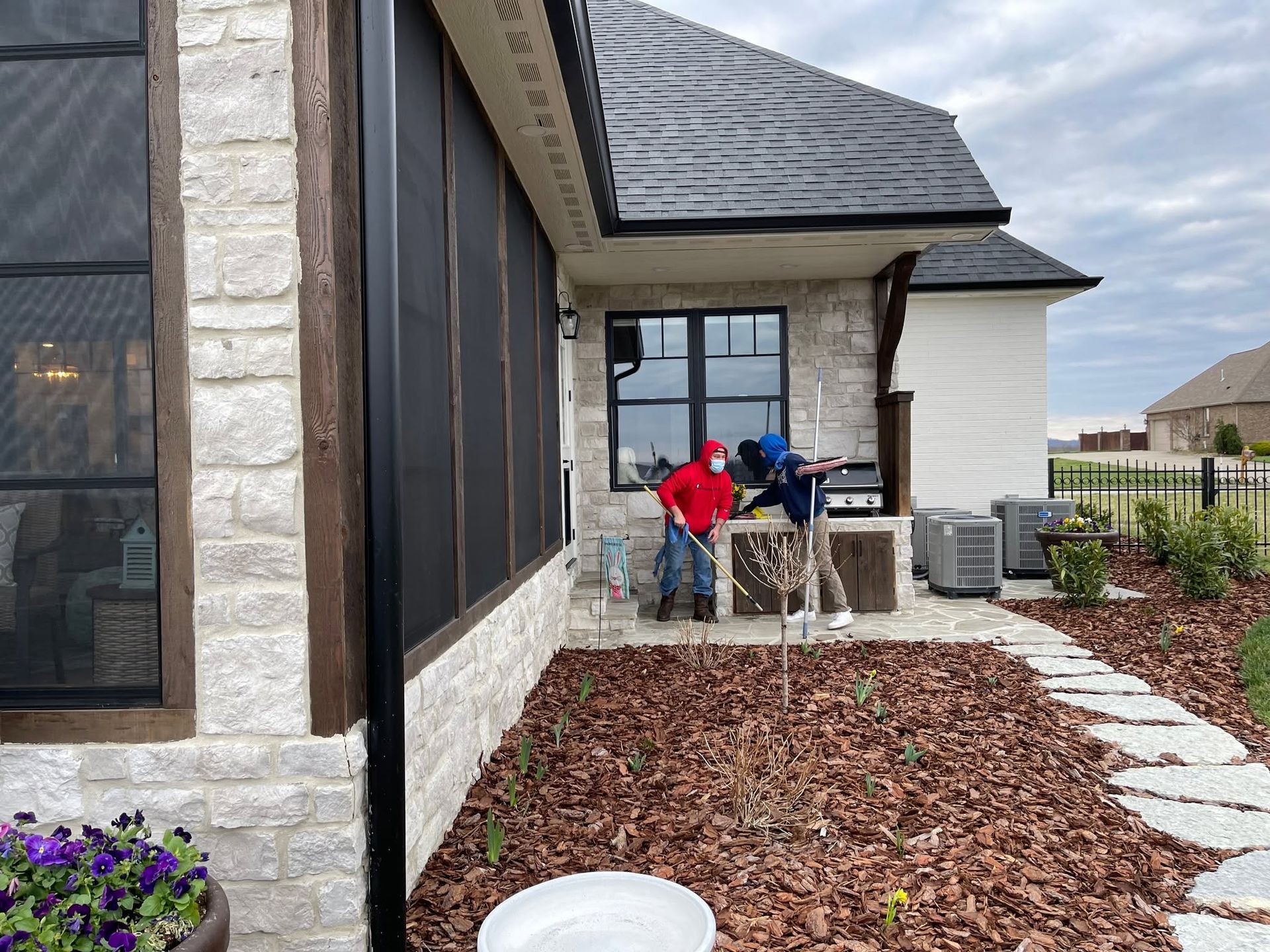 Two people on a patio beside a stone house, with a grill and potted flowers nearby.
