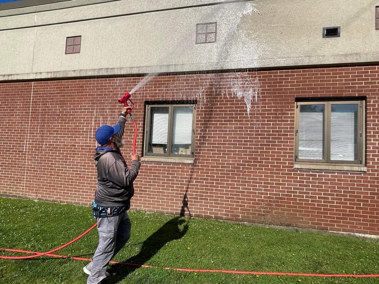 Person pressure-washing a brick wall beside a building, spraying water upward with a red wand.