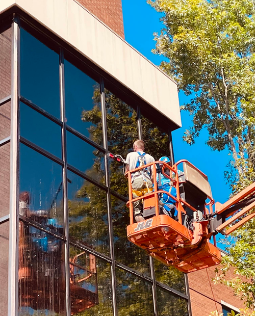 Worker on an orange lift cleaning a modern glass building beside a tree