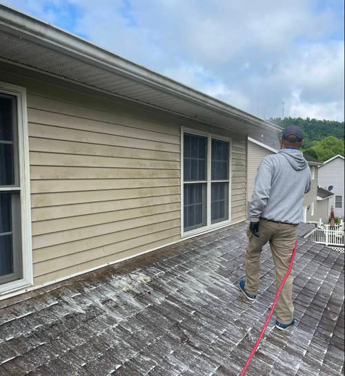 Worker cleaning a roof beside beige siding with a red hose and cloudy sky overhead