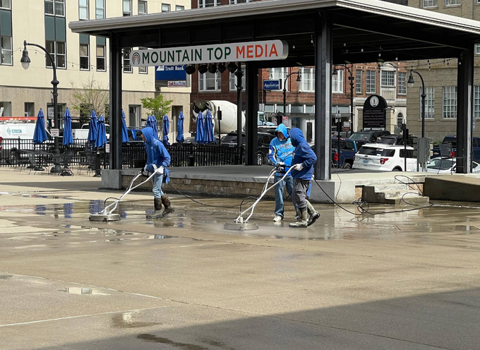 Workers in blue uniforms clean a muddy street in front of a downtown media building.