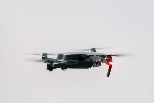 Black quadcopter drone flying against a light gray sky, with a red rear light.