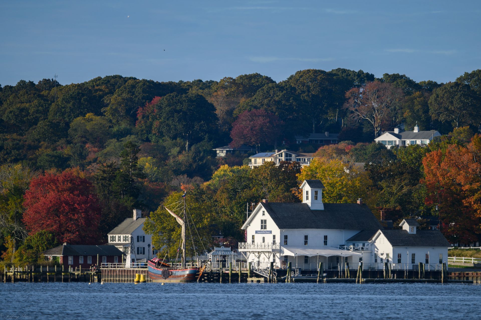 White waterfront buildings and a church on a tree-lined shoreline across calm water under a clear sky