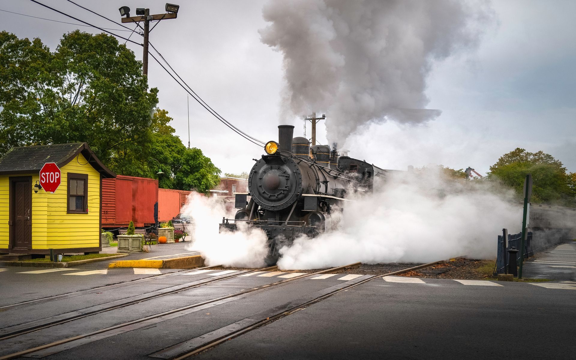 Steam locomotive crossing a road, surrounded by white steam beside a yellow building and trees