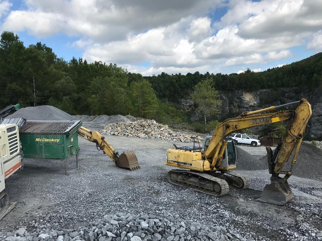 Excavator and machinery at a rock quarry, with pile of rocks and trees in the background.
