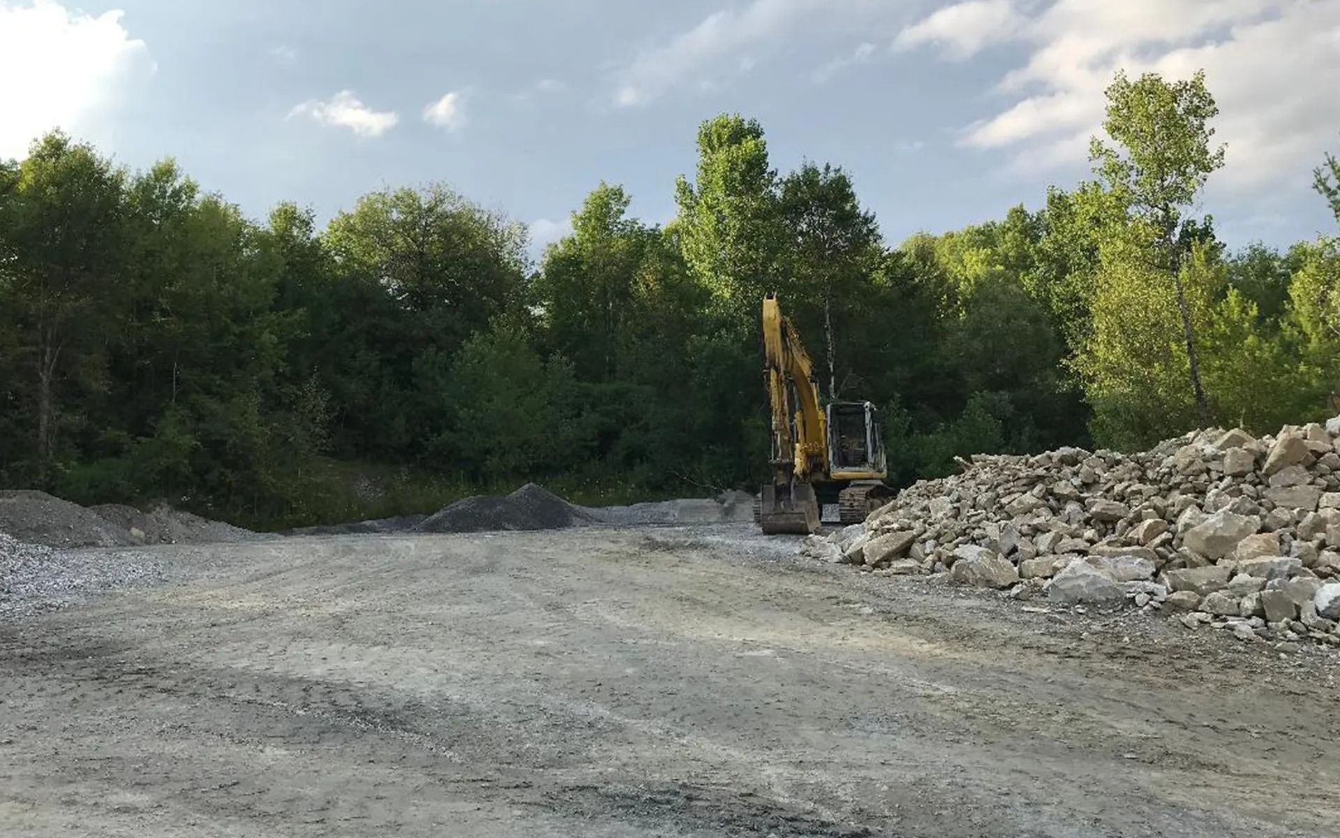 Excavator in a gravel quarry with a pile of rocks and trees in the background under a blue sky.