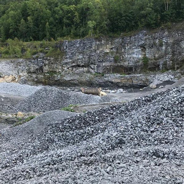 A quarry filled with piles of grey gravel, with a rock face in the background and trees at the top.