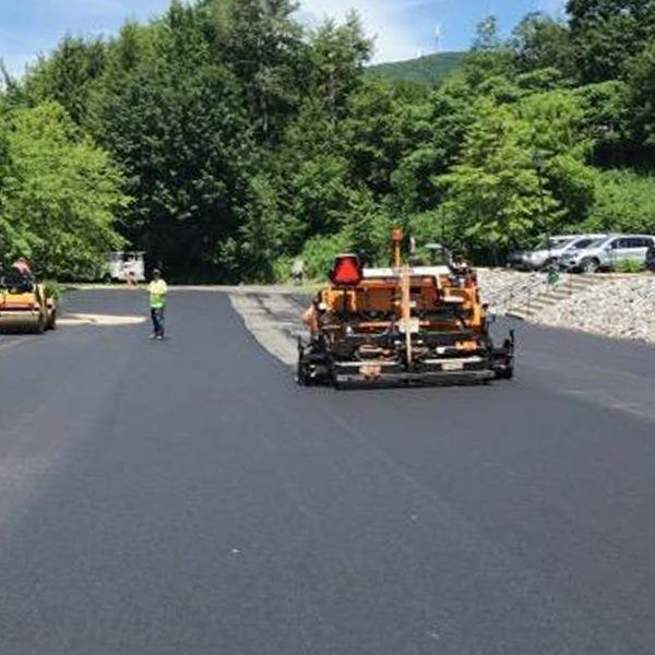 Asphalt paving machine laying blacktop on a road. Man directs the process, surrounded by trees.