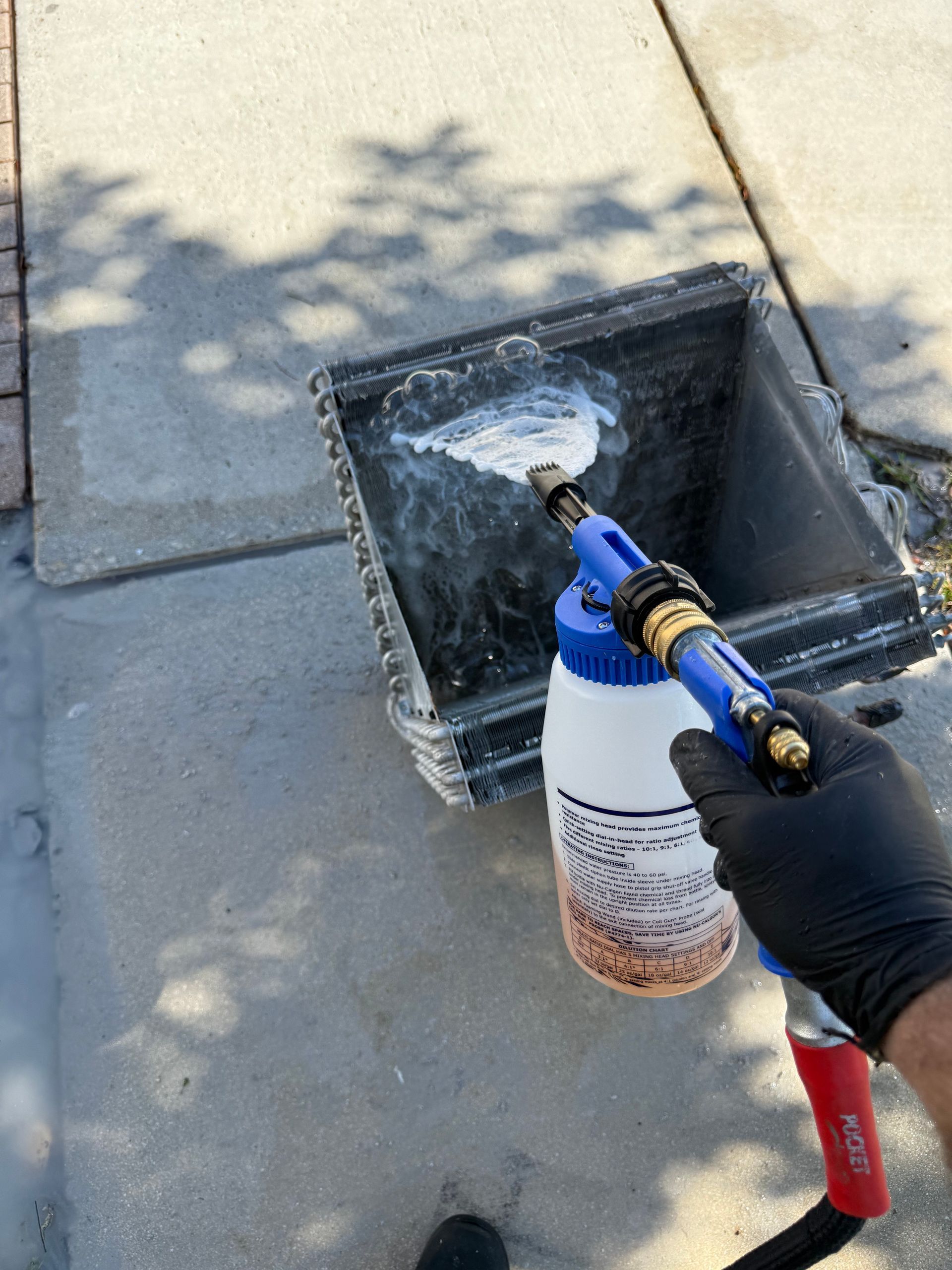 A hand in a black glove uses a spray nozzle to apply white foam cleaner to an air conditioning coil outdoors.