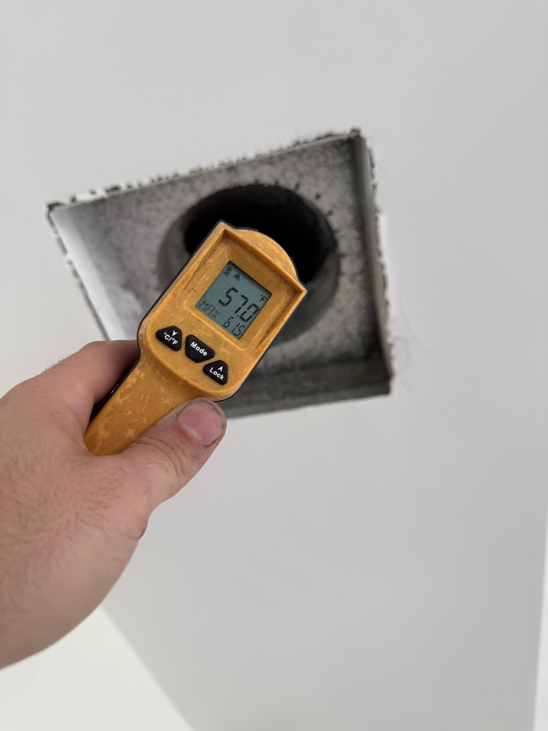 A hand holds a yellow infrared thermometer up to a ceiling vent, showing a temperature reading of 57 degrees.