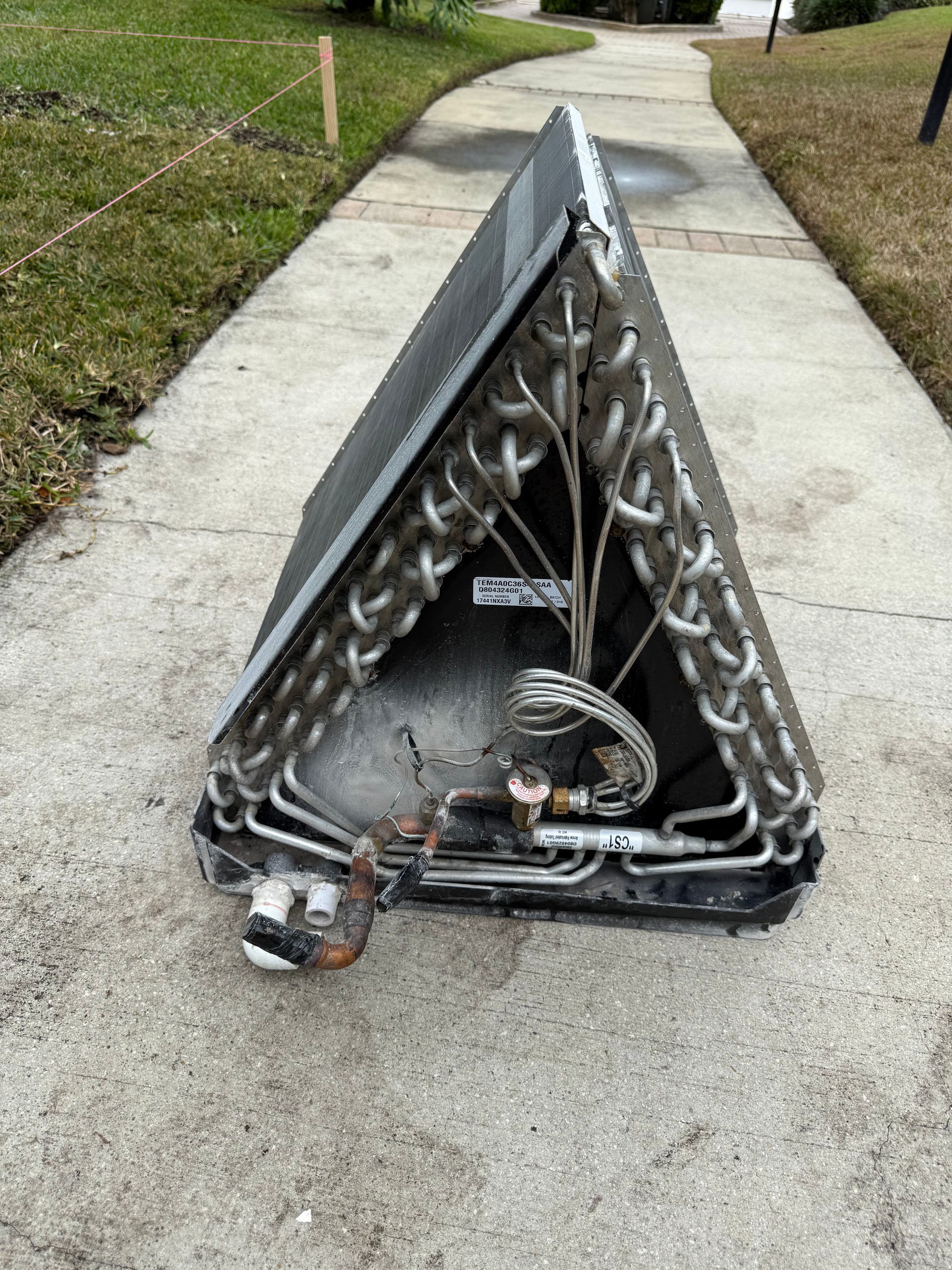 A triangle-shaped HVAC evaporator coil sits on a concrete sidewalk outdoors.