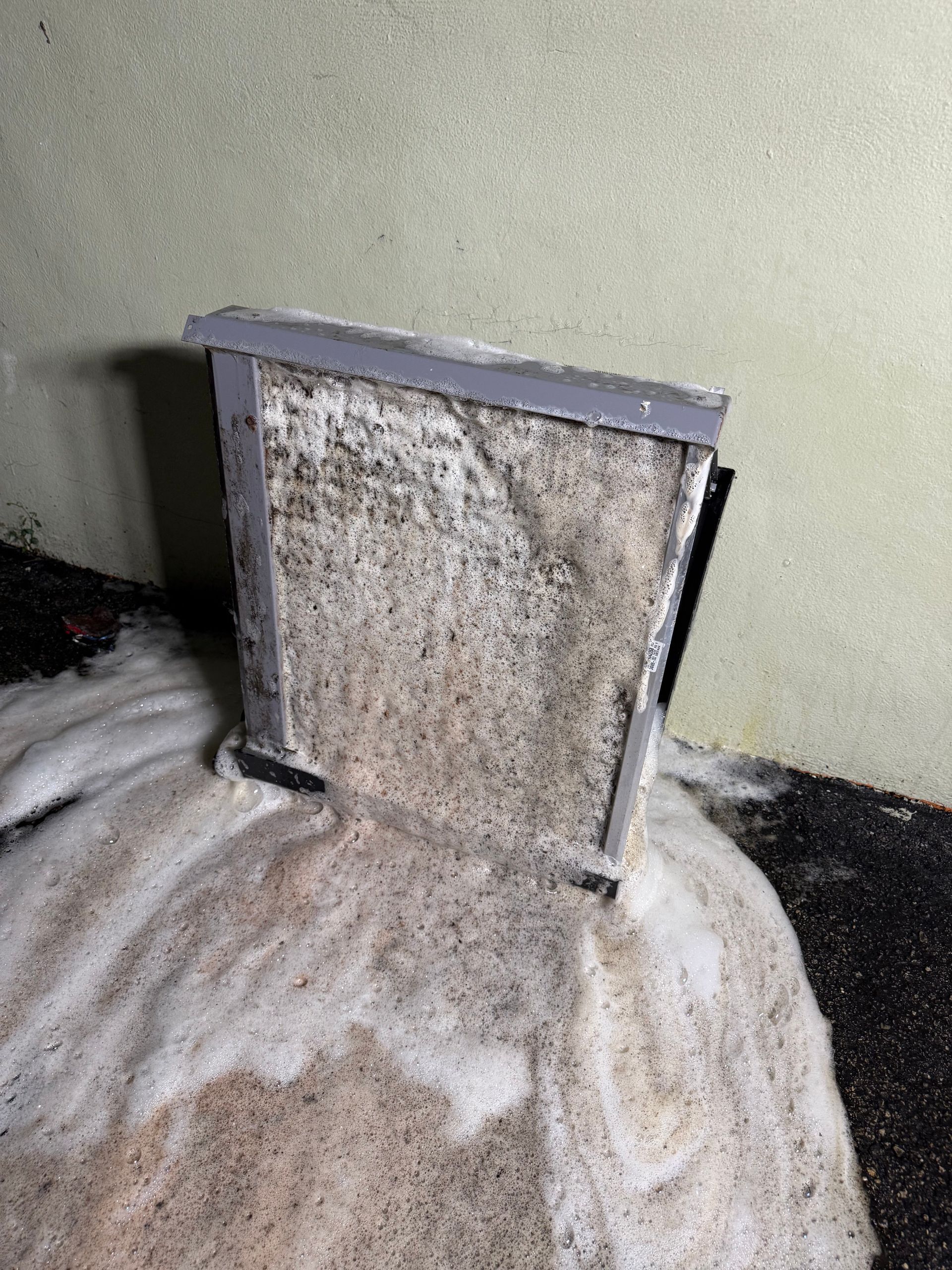 A dirty, used HVAC air filter sits on a surface covered in white cleaning foam against a light-colored wall.