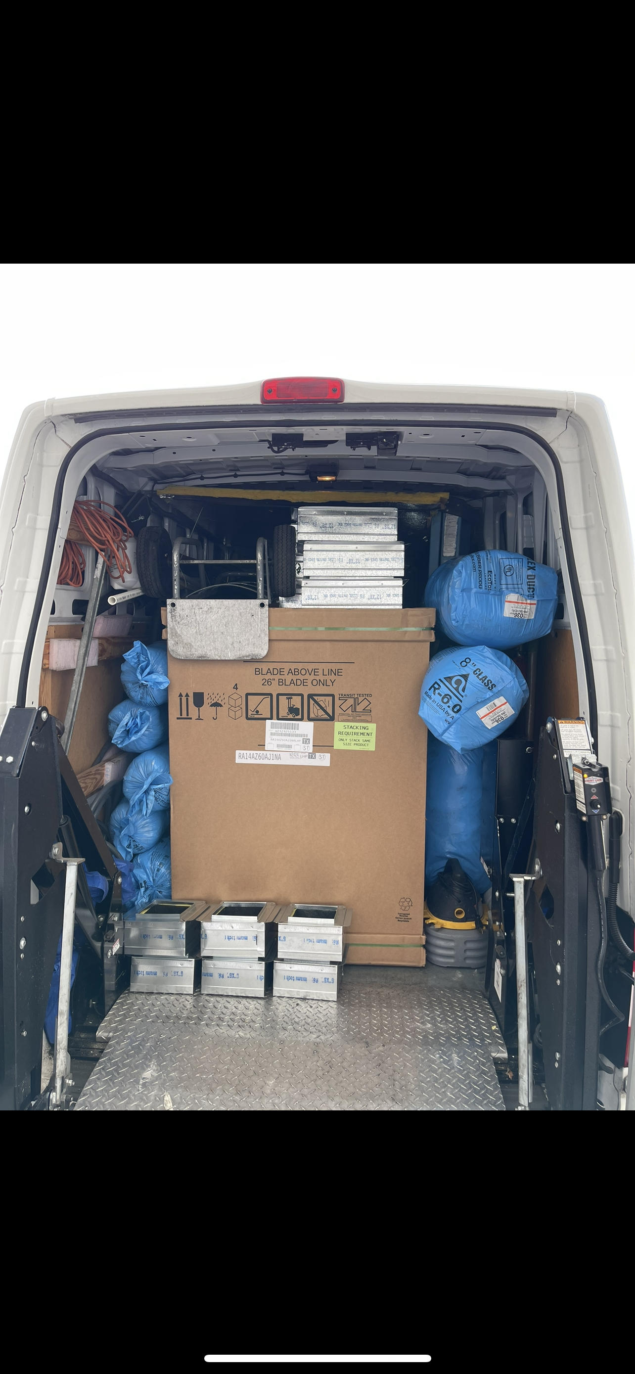 A view inside the back of a delivery van, packed with a large cardboard box, blue bags, and stacked containers.