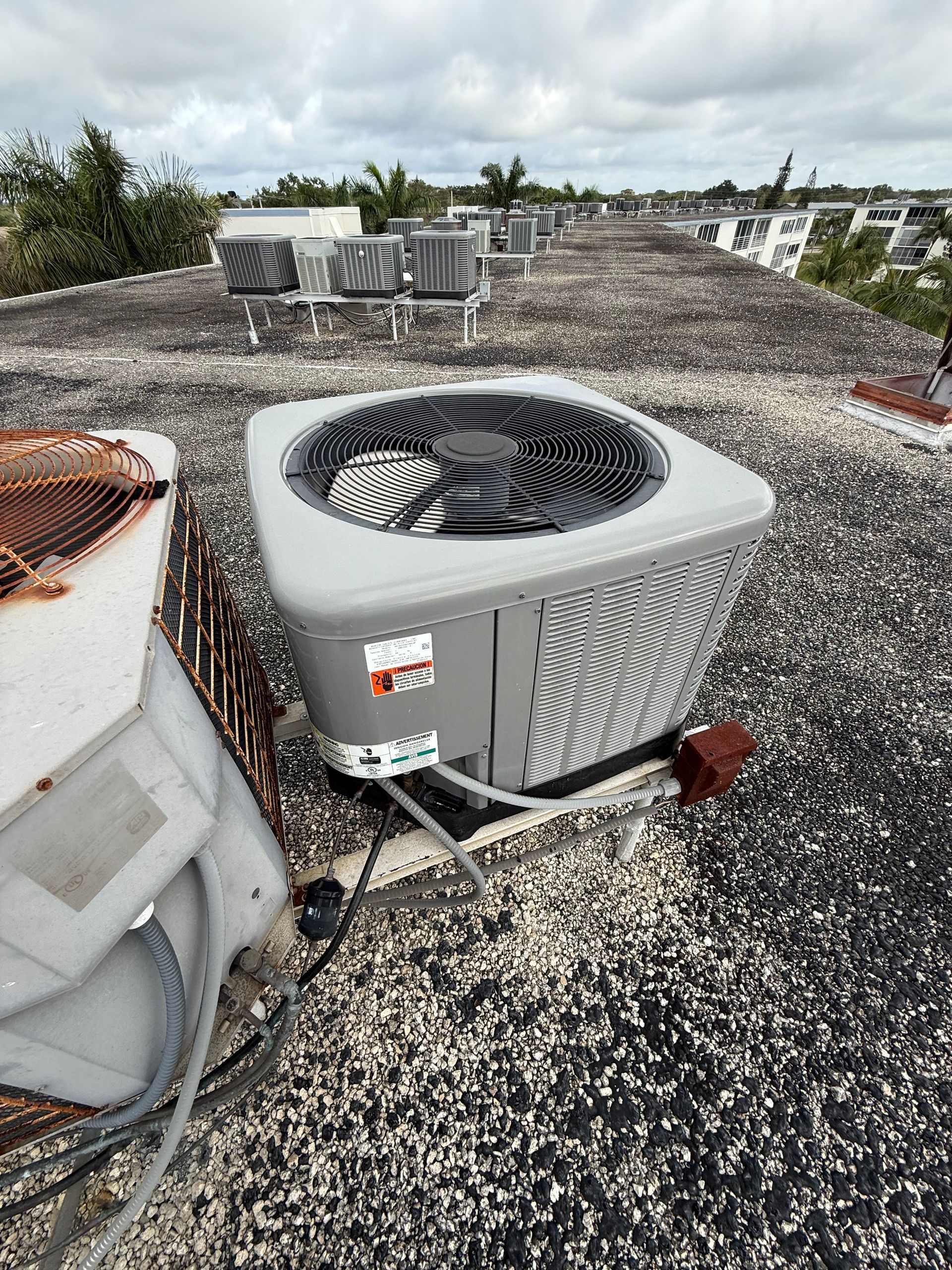 An outdoor HVAC unit on a flat, gravel-covered roof with a partially visible unit next to it under a cloudy sky.