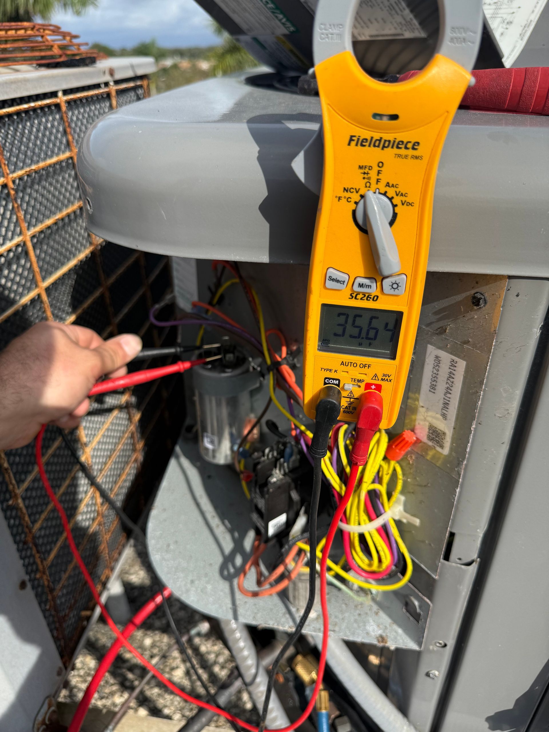 A technician uses a yellow multimeter to test a silver HVAC capacitor, showing a reading of 35.64 on a unit's control panel.