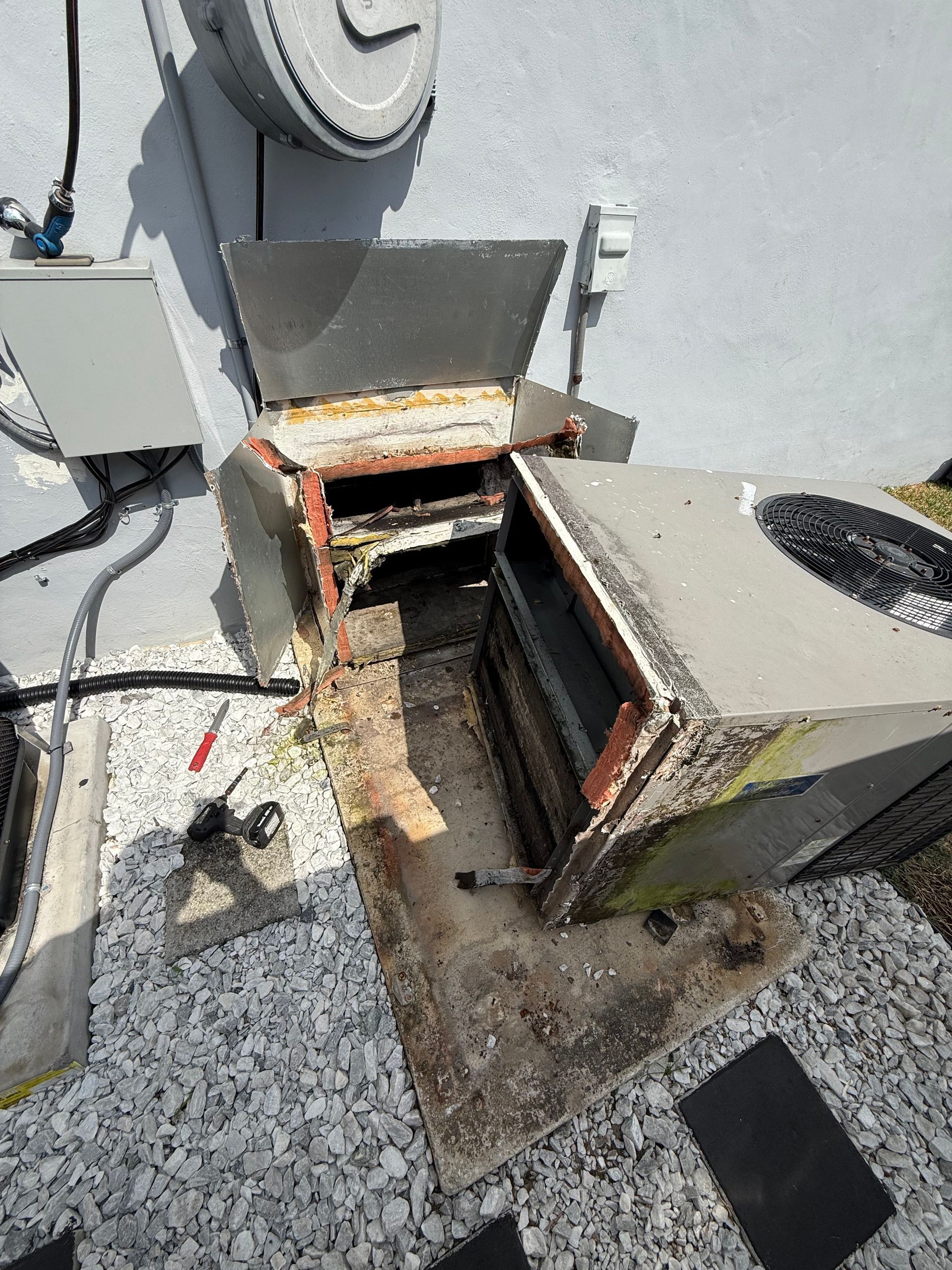 Outdoor HVAC unit with open, rusted access panels exposing internal components on a stone-covered ground surface.