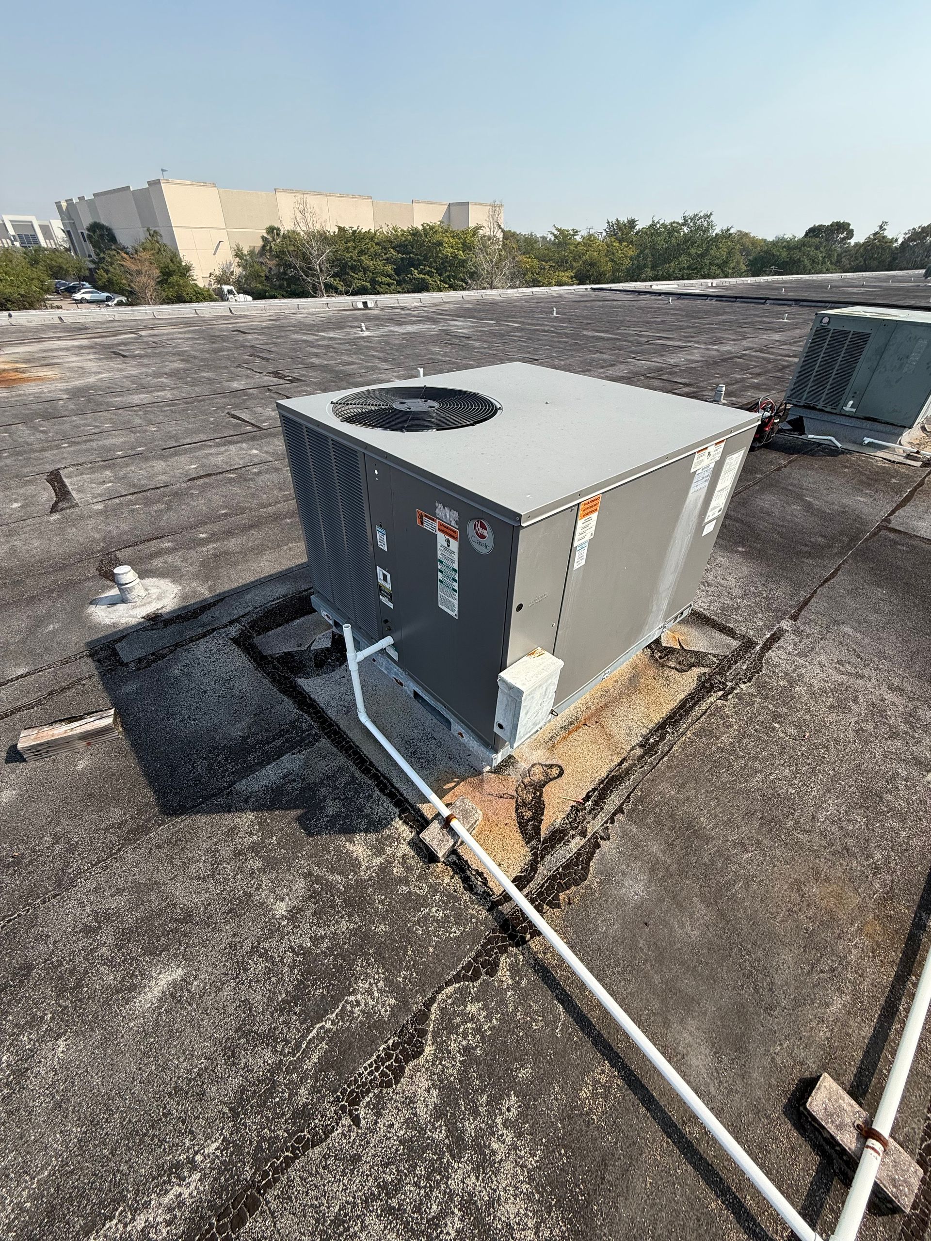 A gray HVAC unit sits on a flat, dark gravel rooftop with white PVC piping running across the surface.