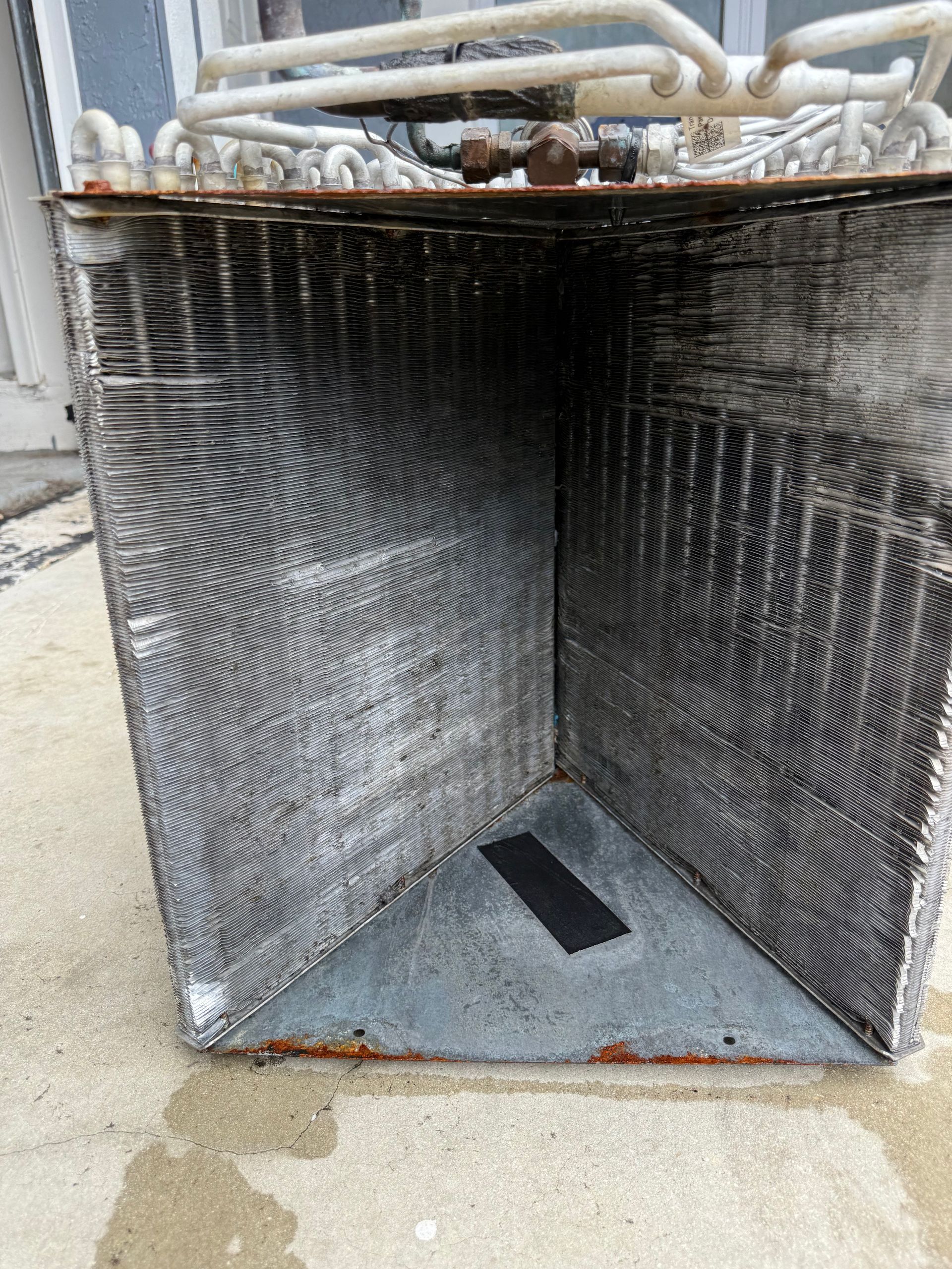 A dirty, V-shaped HVAC evaporator coil sits on a concrete surface, showing accumulated dust and debris on the fins.