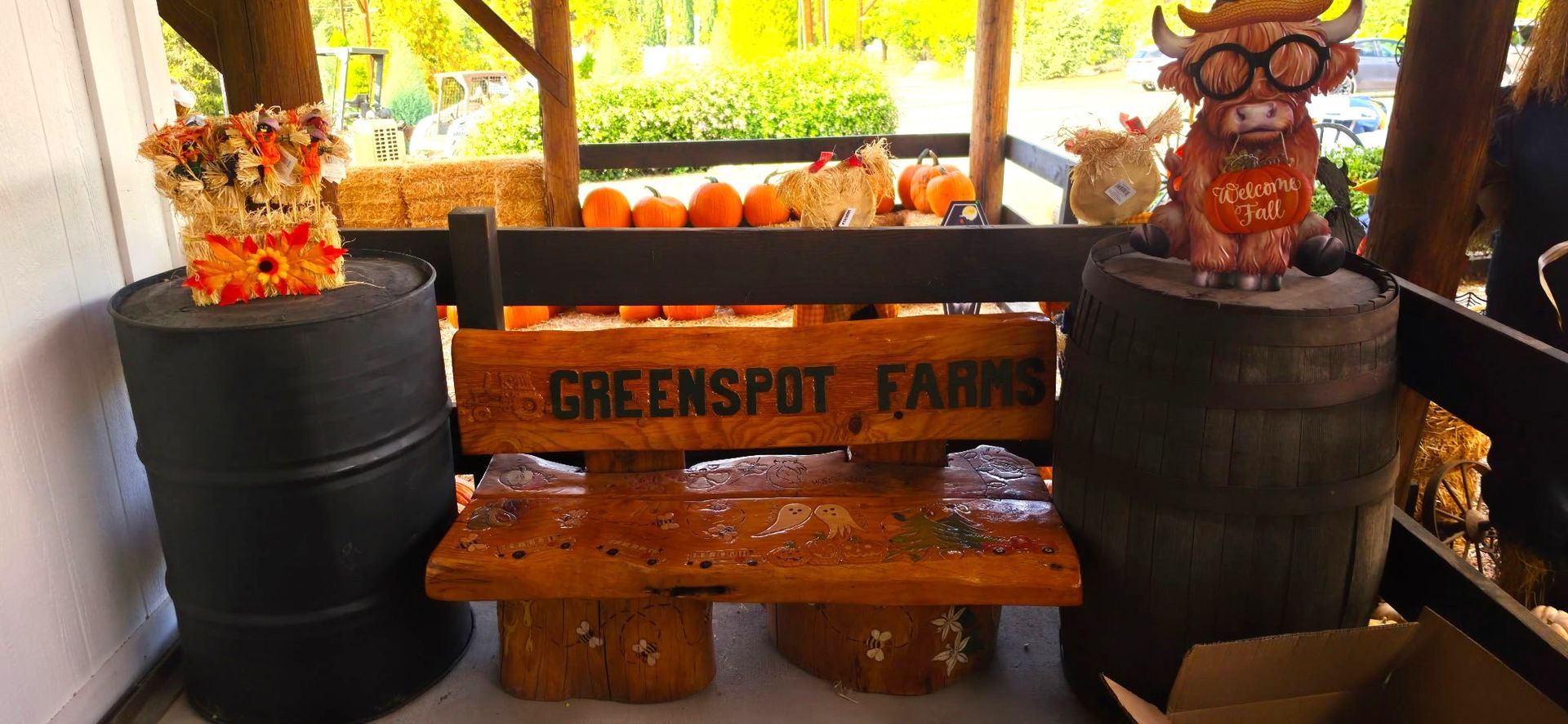 Bench labeled “Greenspot Farms” between two black barrels topped with autumn decor and a small owl figure