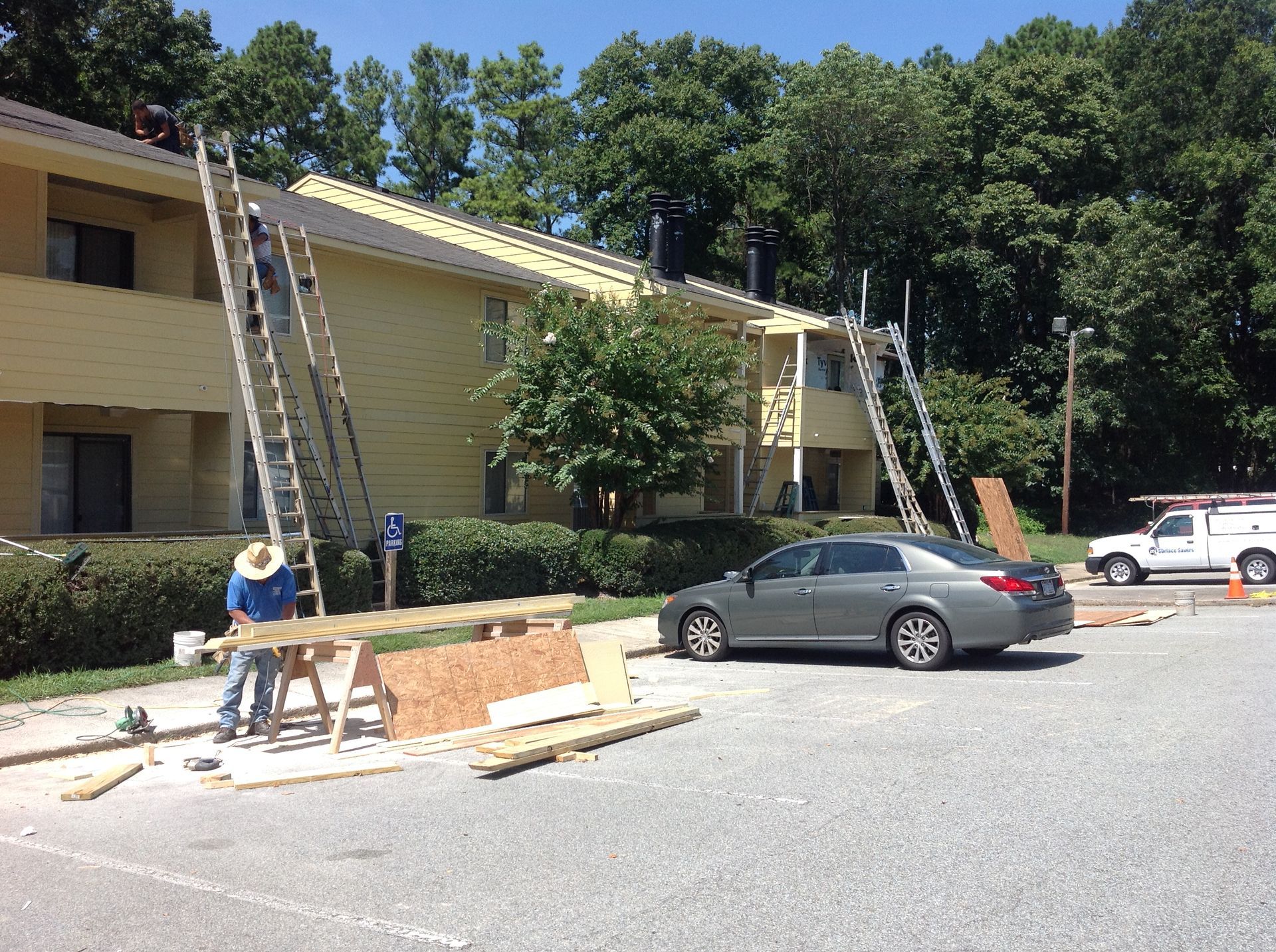 Workers on ladders repairing a light yellow apartment building roof on a sunny day. A car and truck are nearby.
