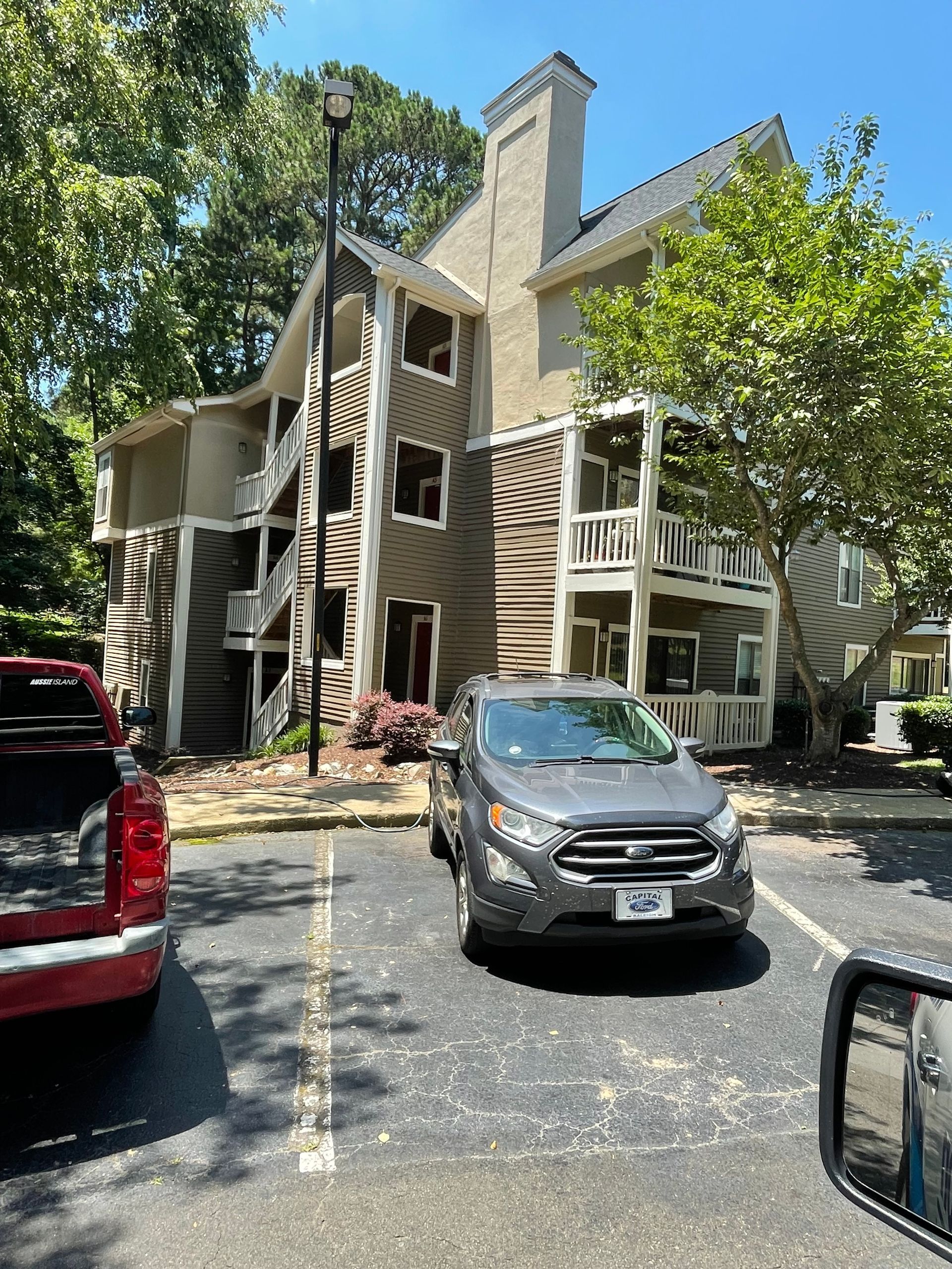 Exterior of a beige apartment building with external stairways. A gray SUV is parked in front, with a red pickup on the left.