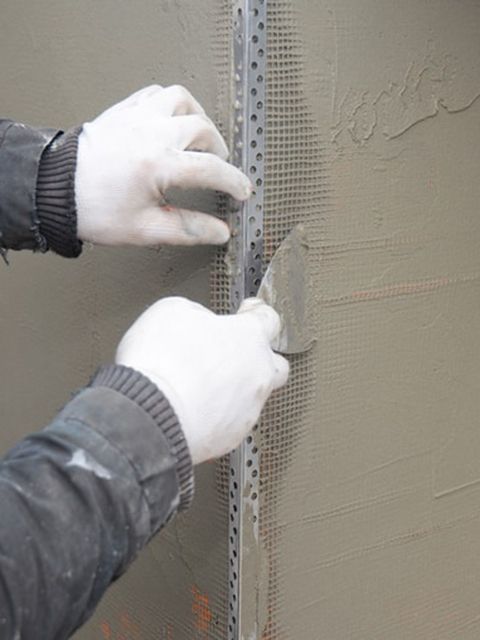 Hands in gloves smoothing plaster with a trowel along a metal corner bead on a wall.