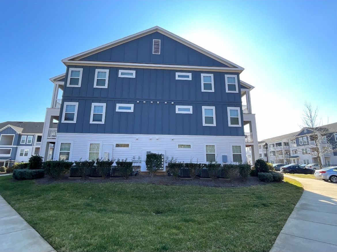Blue and white multi-story apartment building under a bright blue sky. Green grass and sidewalk in foreground.
