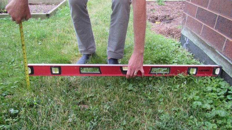 Person measuring grass with a level and tape measure near a brick wall.