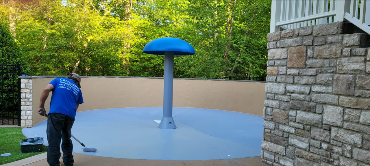 Man painting a blue splash pad with a mushroom-shaped fountain. Green trees in the background, stone wall to the right.