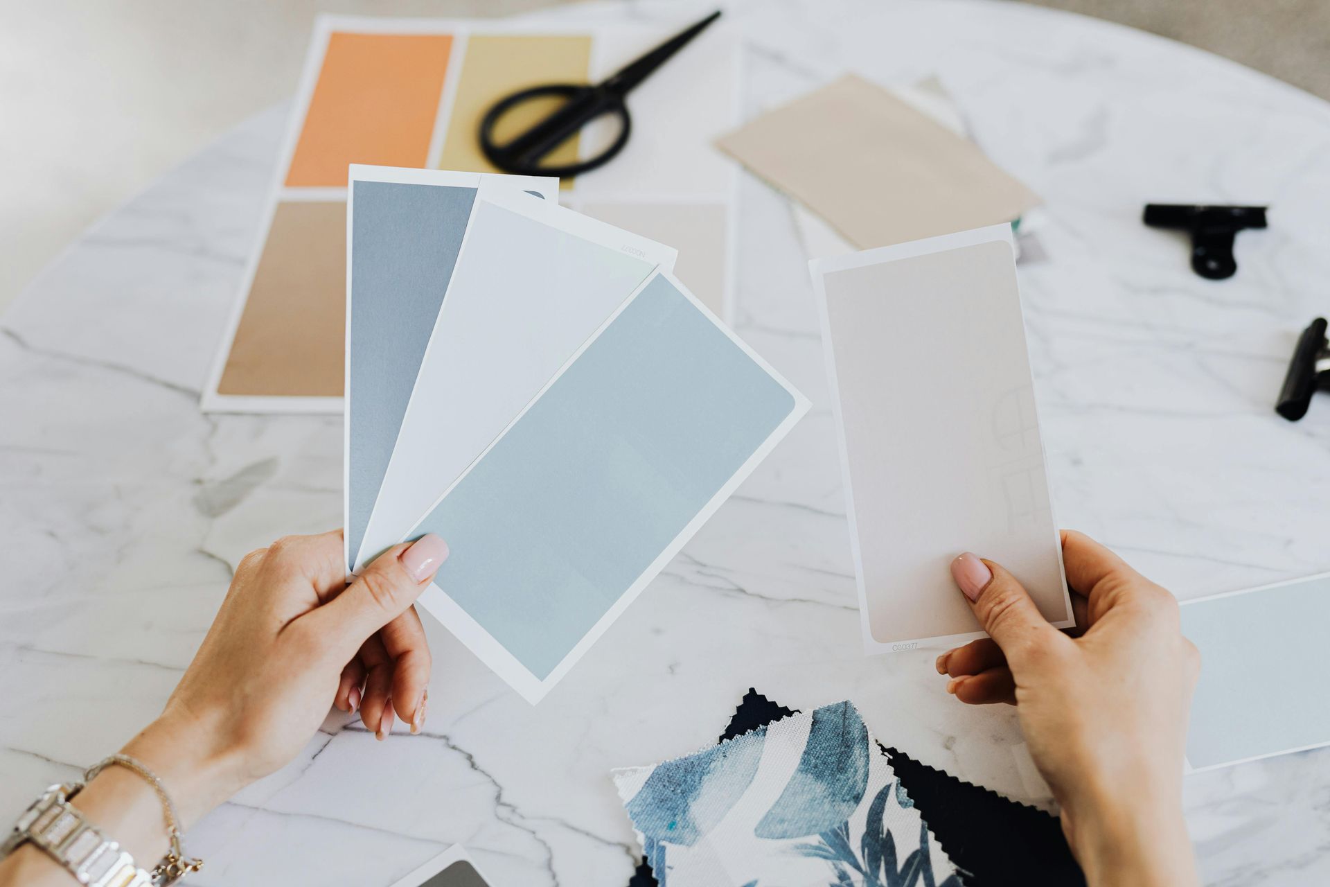 Hands holding paint swatches on a table with other color samples, scissors, and a clip.