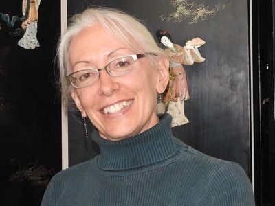 A woman wearing glasses and a turtleneck is smiling in front of a painting.