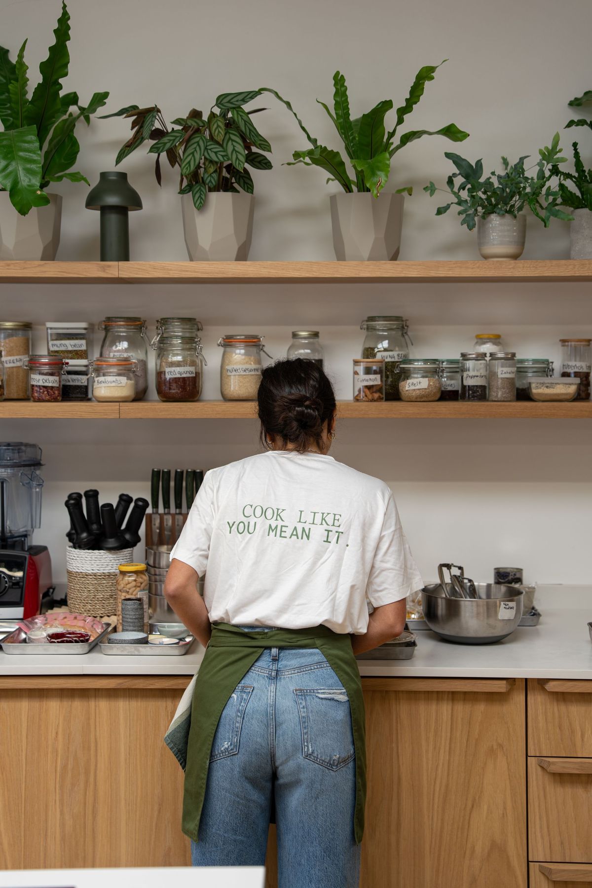 Academy student in a Cook Like You Mean It t-shirt and green apron at the studio kitchen shelves, Bettina’s Kitchen by REGO Creative