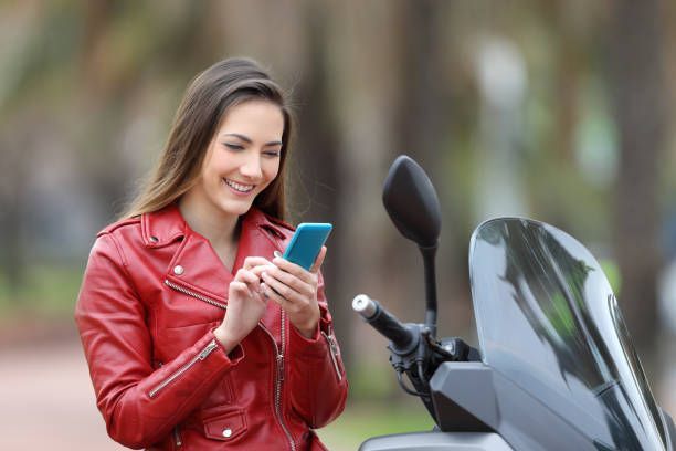 A woman in a red leather jacket is standing next to a motorcycle and looking at her cell phone.