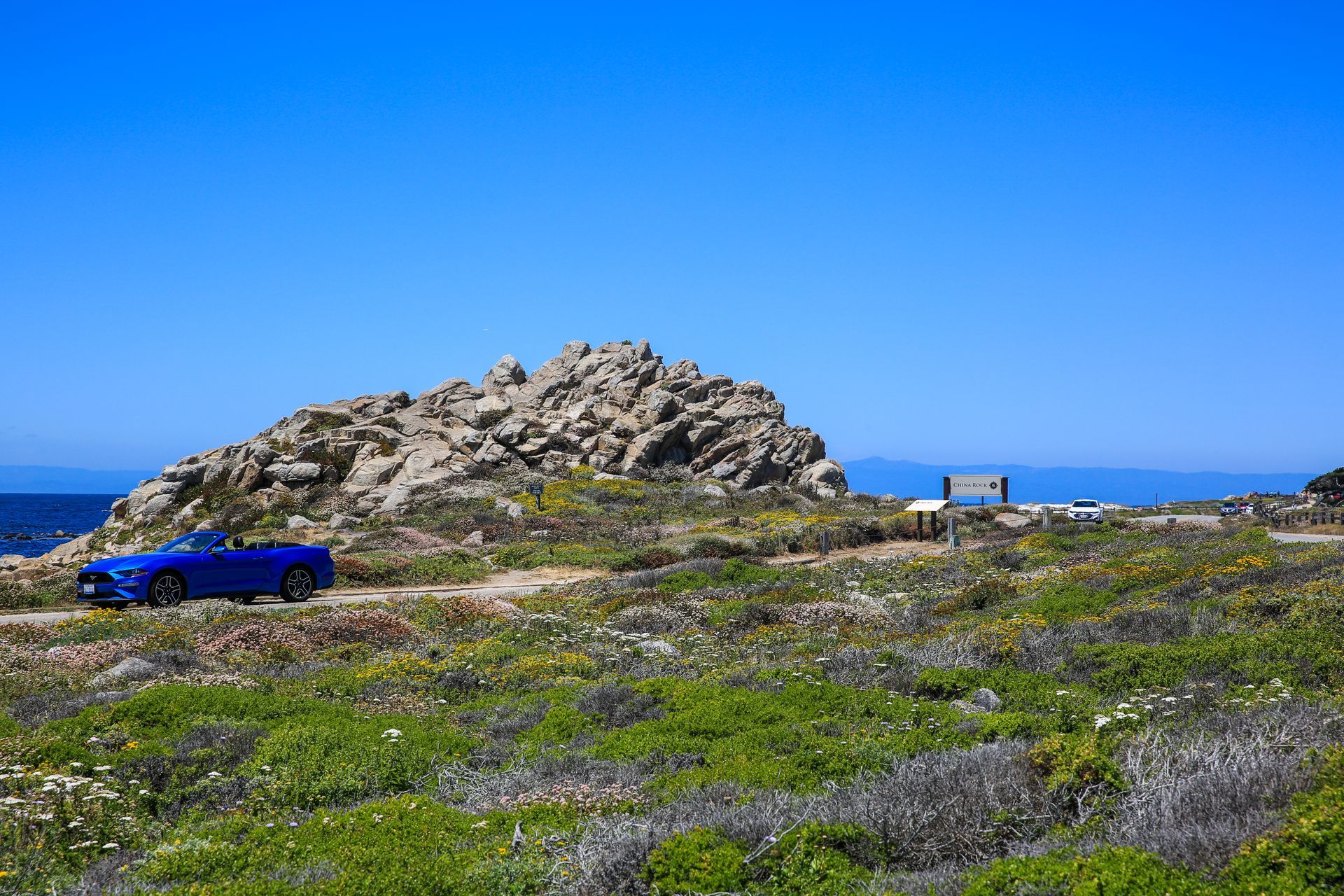 Blue car near rocky coastal outcrop with wildflowers under a clear blue sky