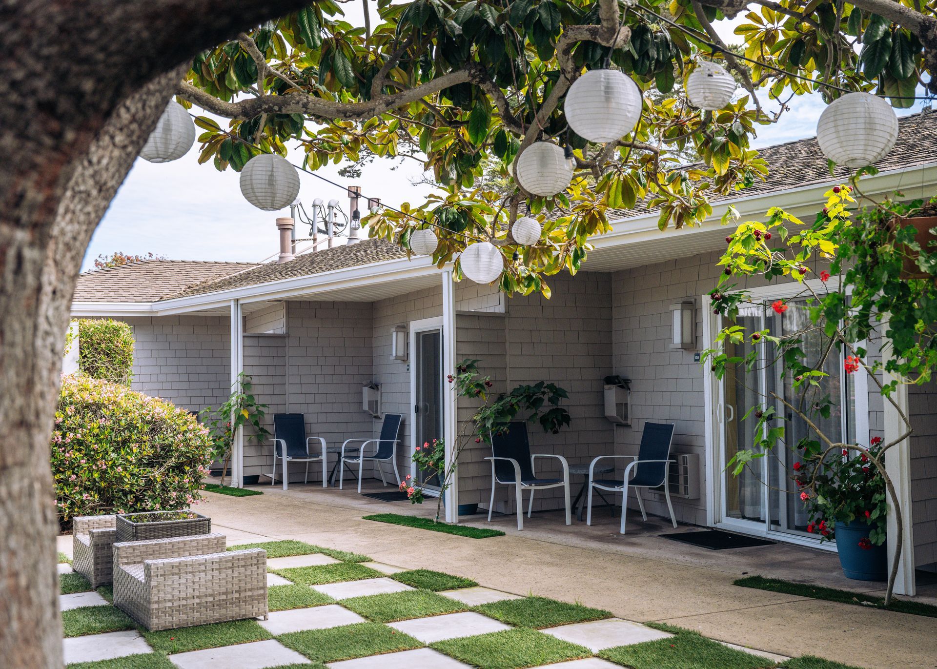 Patio with white chairs, hanging lanterns, and a tiled walkway beside a house garden