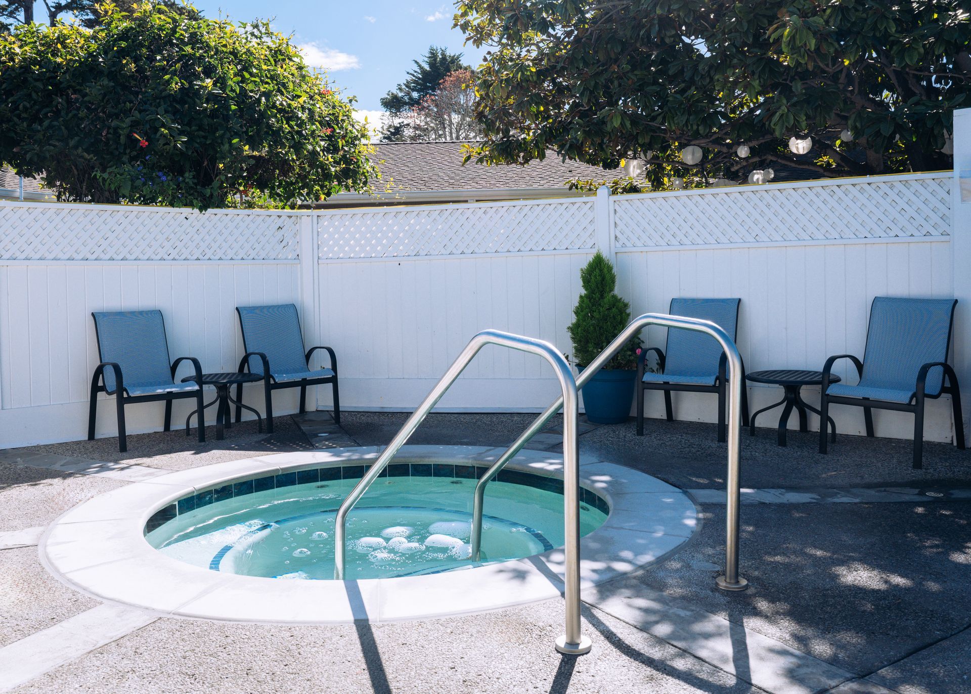 Small backyard spa pool with metal handrails and blue chairs against a white fence