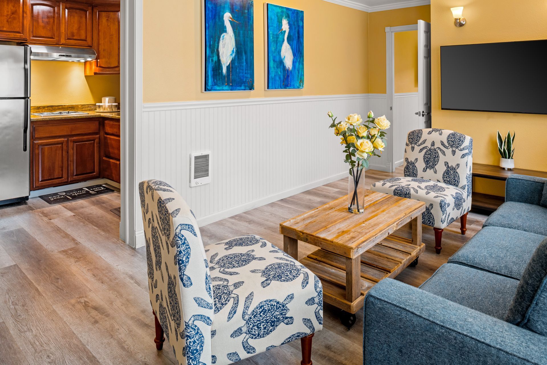 Cozy living room with blue sofa, floral chairs, wooden coffee table, wall art, and open kitchen view